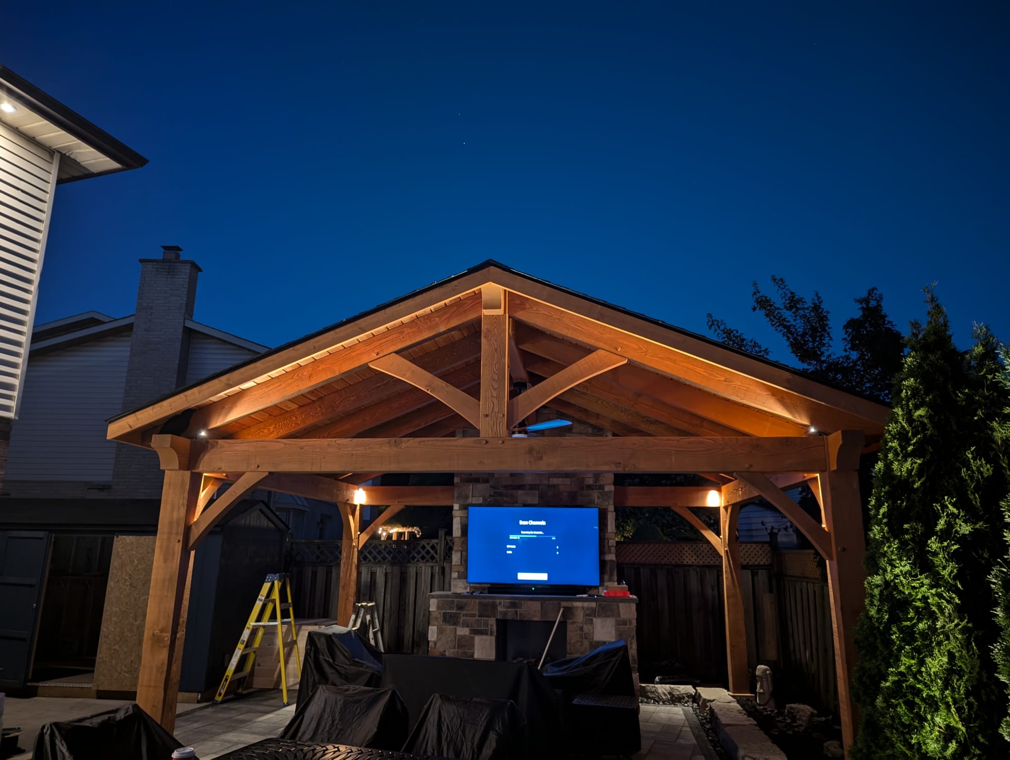 A wooden pavilion at night with outdoor lighting, a ladder, and a covered patio area. A TV displays a setup screen.