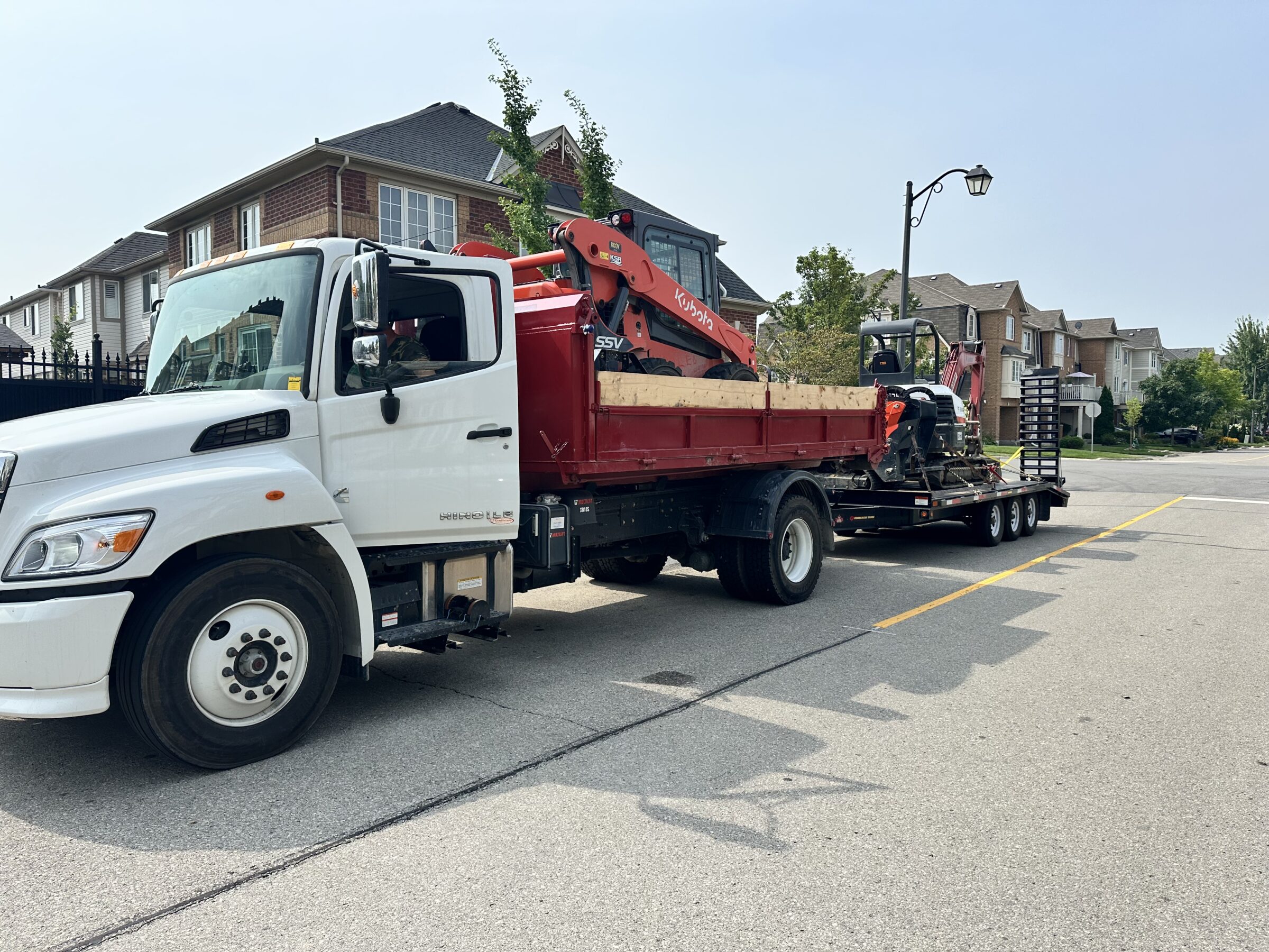A white truck hauls construction equipment in a suburban neighborhood. Brick houses and a streetlamp line the residential street.