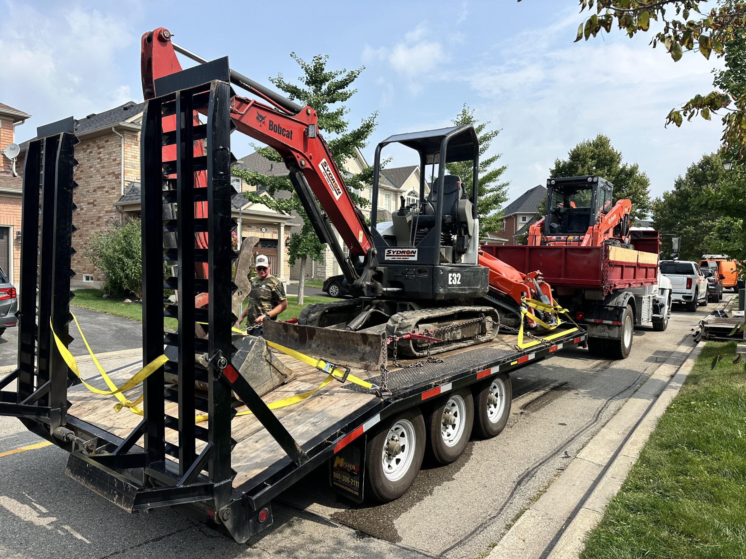 A small excavator loaded on a trailer in a residential neighborhood. A person stands nearby. Trees and parked vehicles line the street.