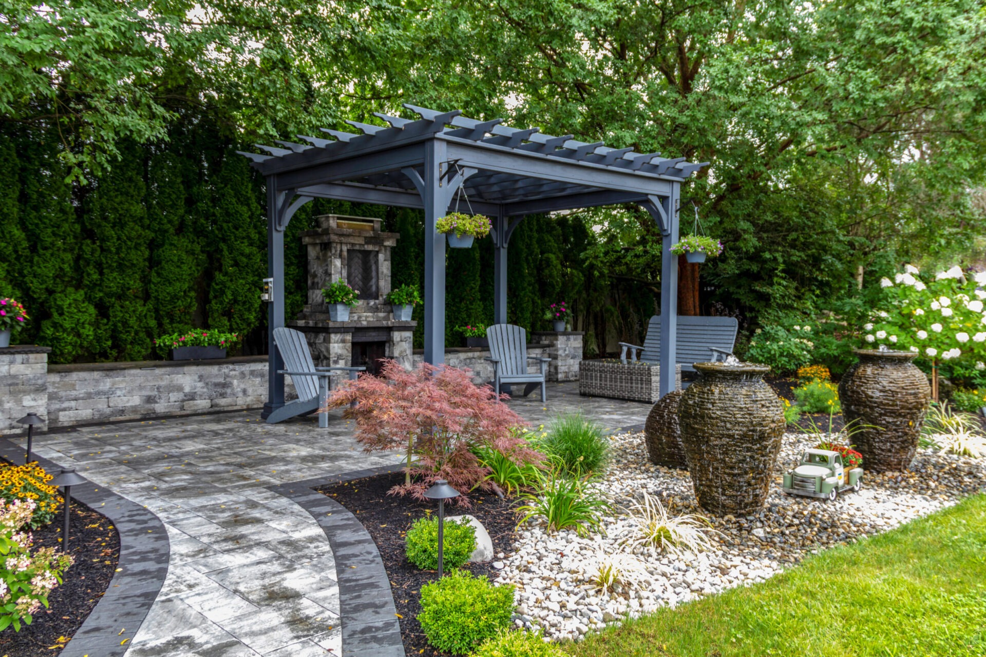 Large gray pergola on pavers with stone fireplace and waterfall urns for backyard oasis landscape architecture