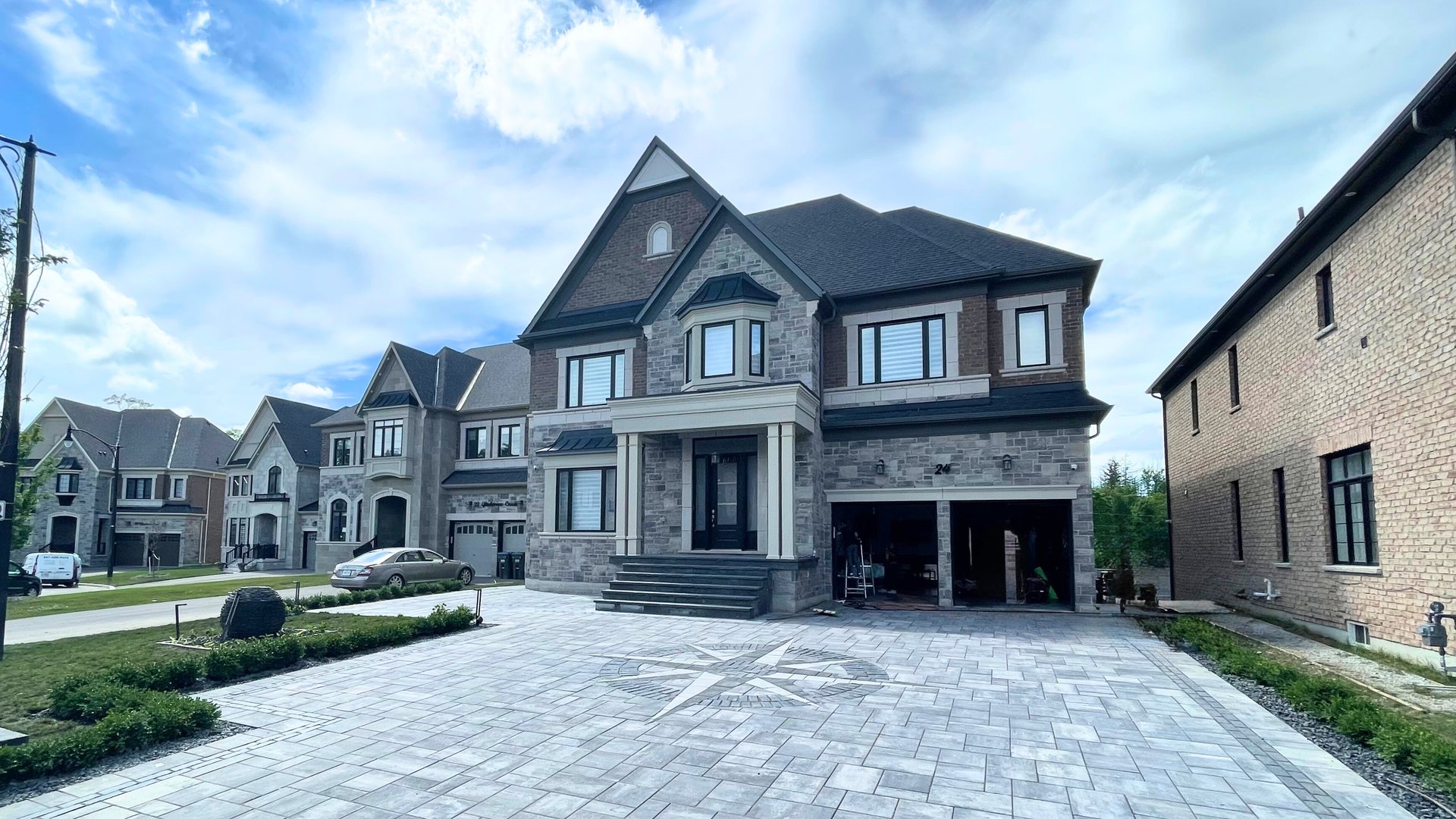 Modern two-storey stone and brick home with a wide front entrance, large custom interlock driveway featuring a compass inlay, and a neatly landscaped front yard in a suburban neighbourhood.