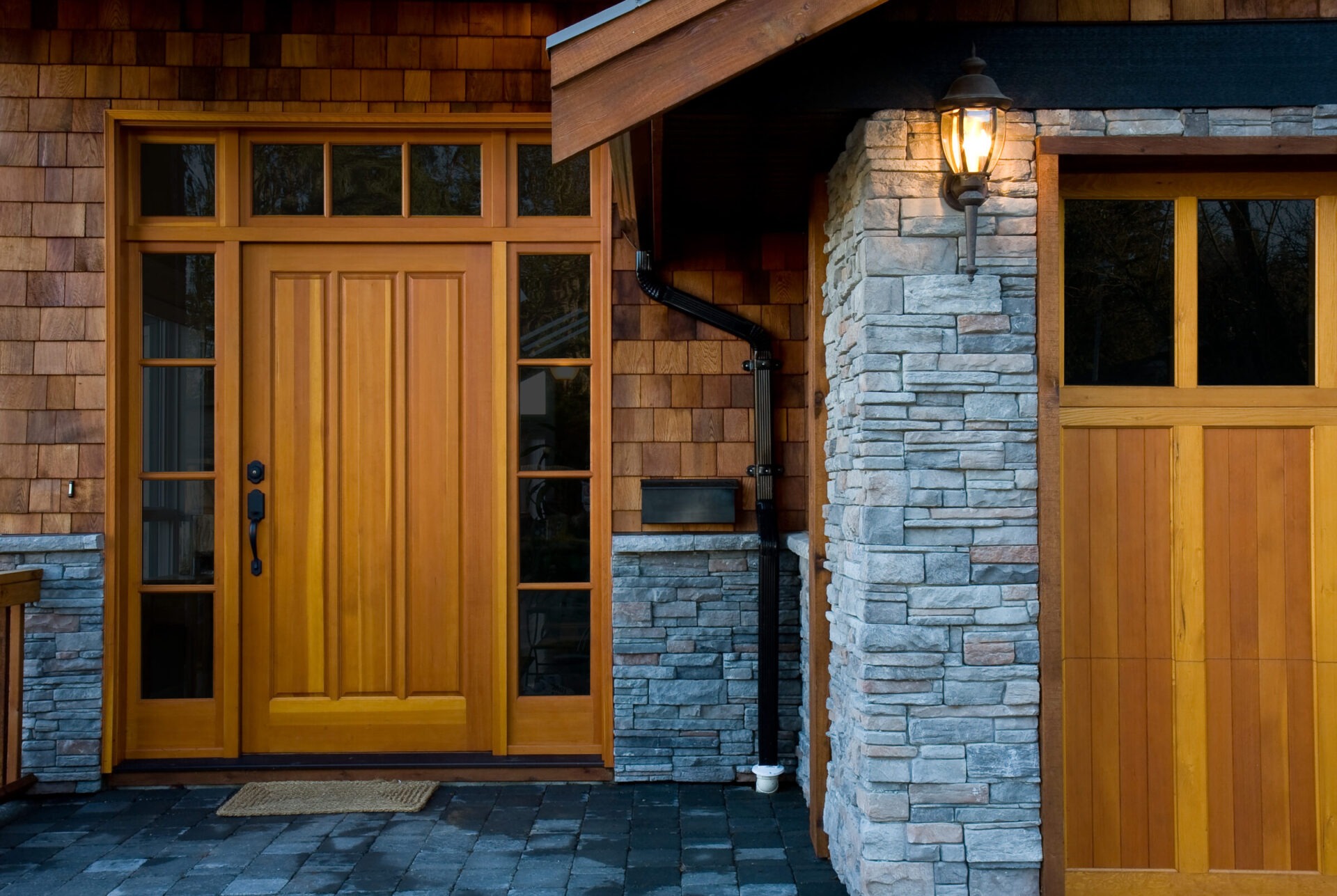Wooden front entry door and garage with natural stone accents and warm exterior lighting on a craftsman-style home.