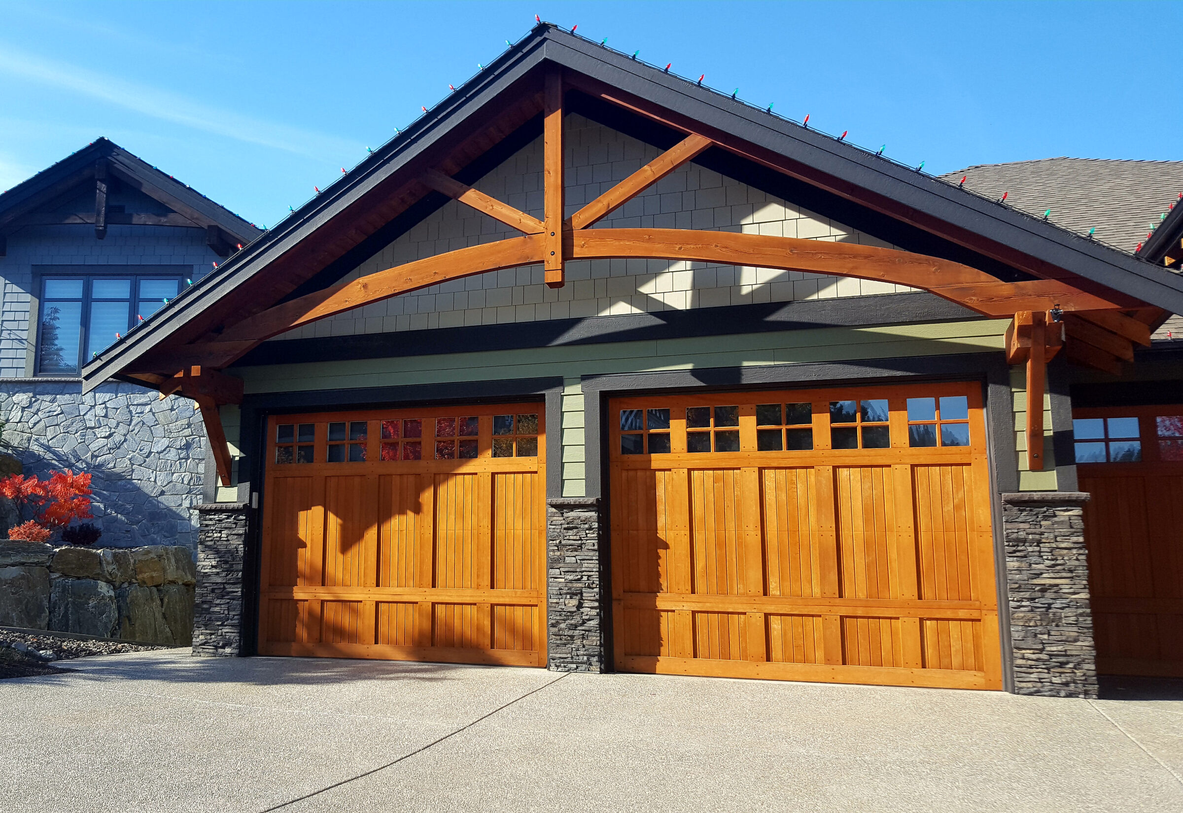 Double wooden garage doors beneath a gabled roof with exposed timber beams and stone pillars on a craftsman-style home exterior.