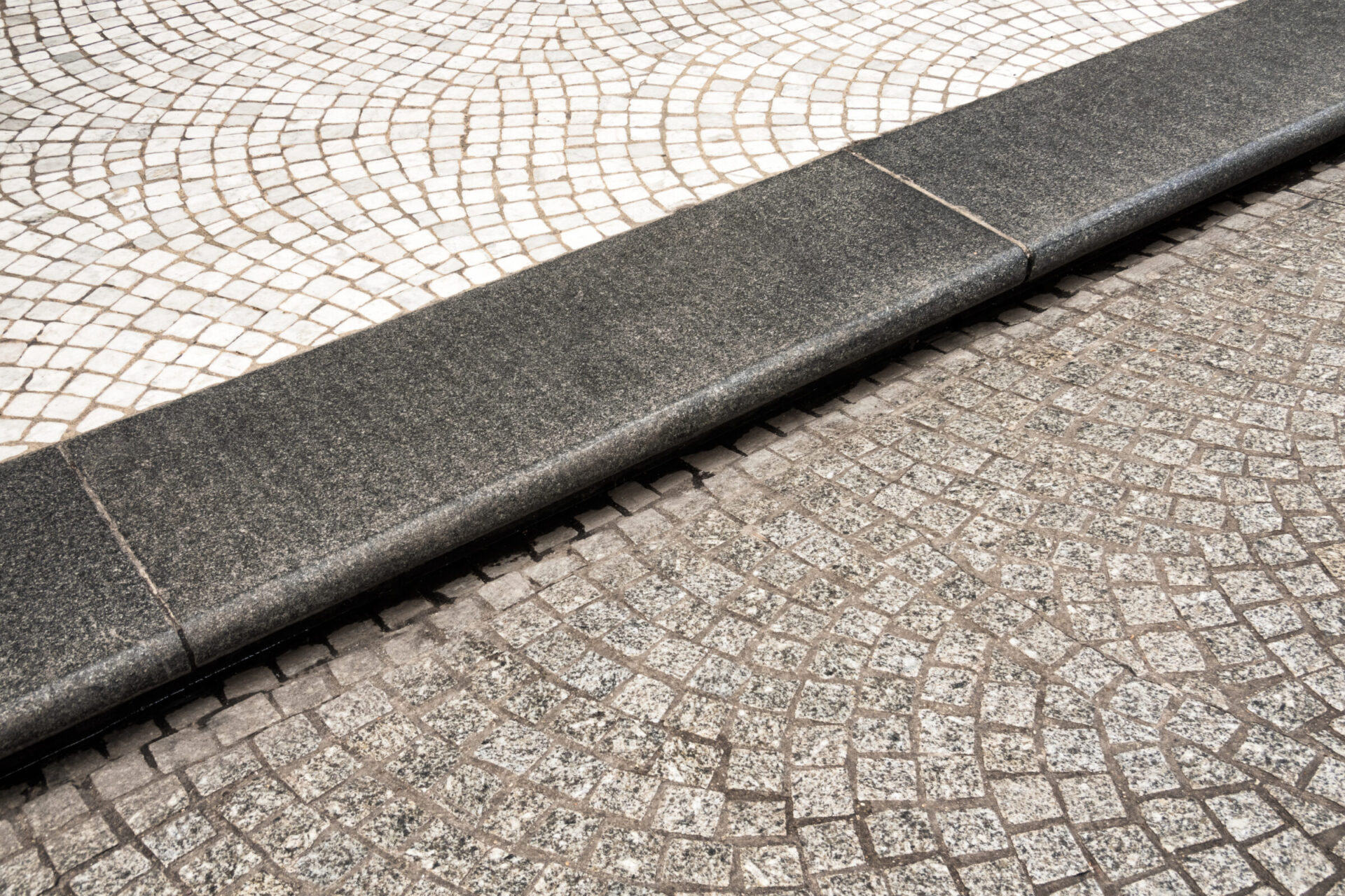 Close-up of a curved granite interlock pattern with contrasting light and dark cobblestones, separated by a smooth dark stone curb, showcasing precision stonework in driveway or walkway design.