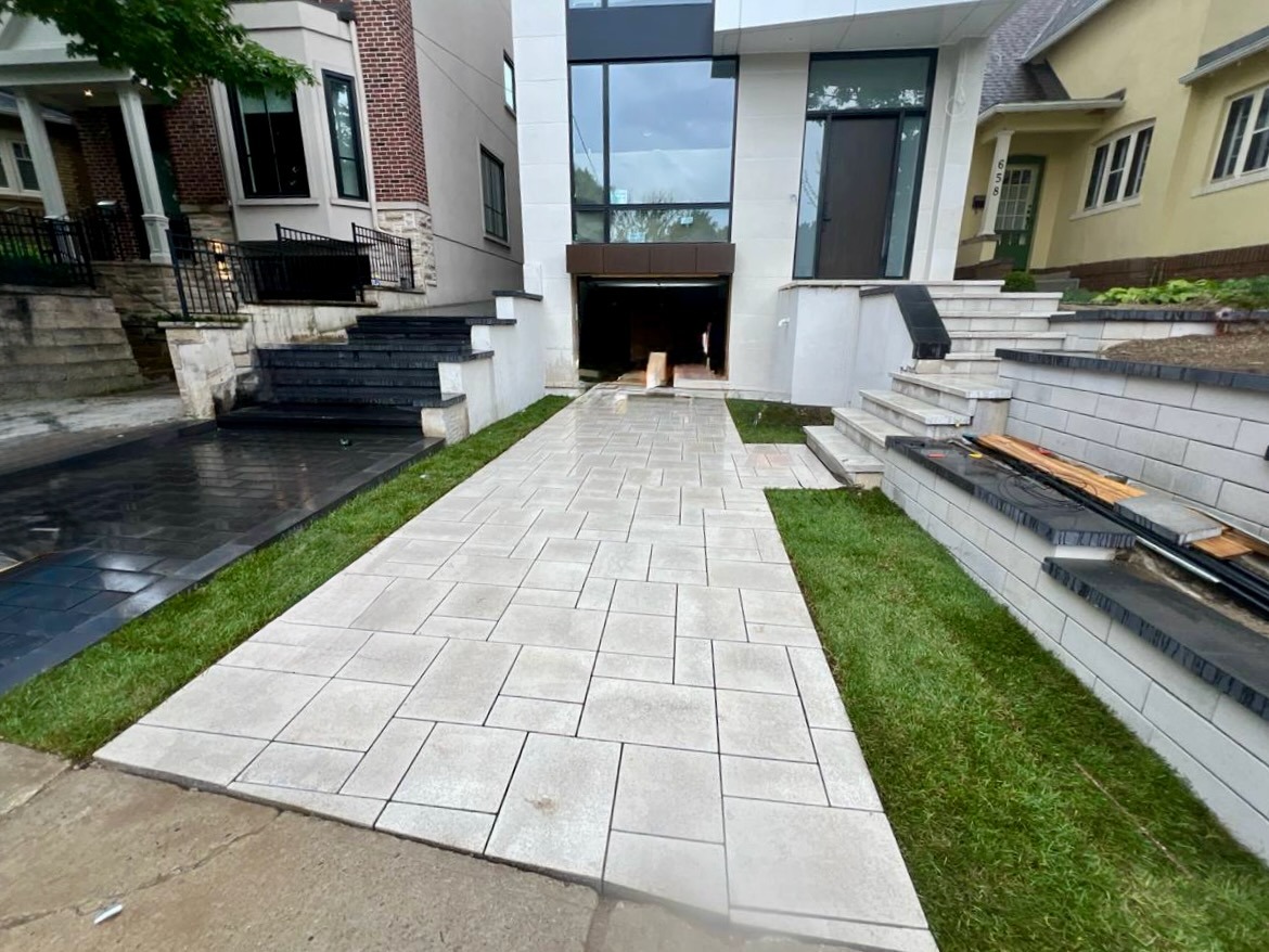 Freshly installed light gray interlock driveway leading to a modern home with large glass windows, flanked by tiered retaining walls, clean steps, and well-manicured grass edging.